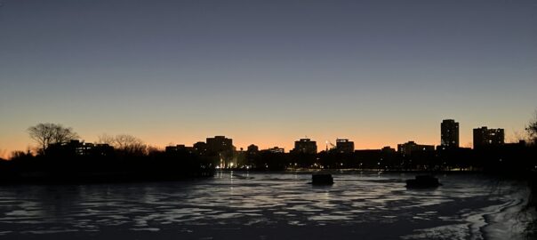 an orange glow of pre dawn on the horizon reflects on the frozen surface of the Rideau River, silhouetting the builidngs and trees of the skyline.