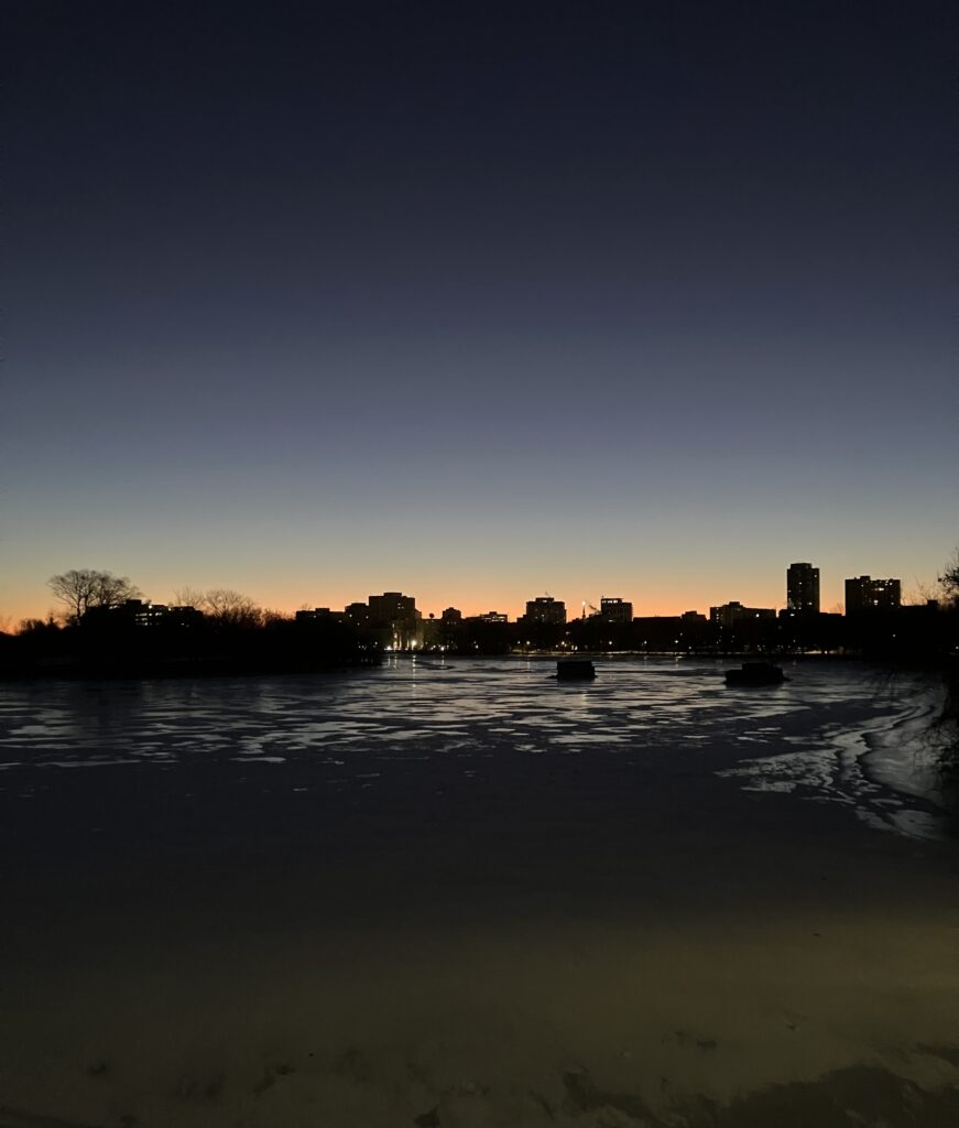 an orange glow of pre dawn on the horizon reflects on the frozen surface of the Rideau River, silhouetting the builidngs and trees of the skyline.